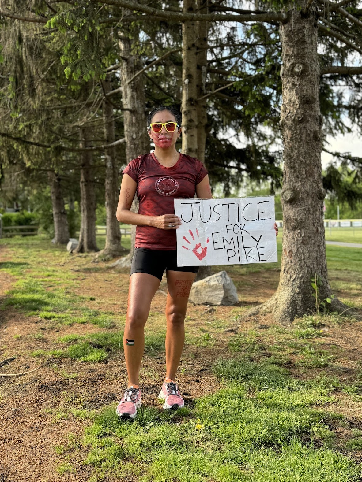 A woman standing outdoors next to trees, holding a sign that reads "JUSTICE FOR EMILY PIKE" with a handprint symbol. She wears sunglasses and athletic attire.